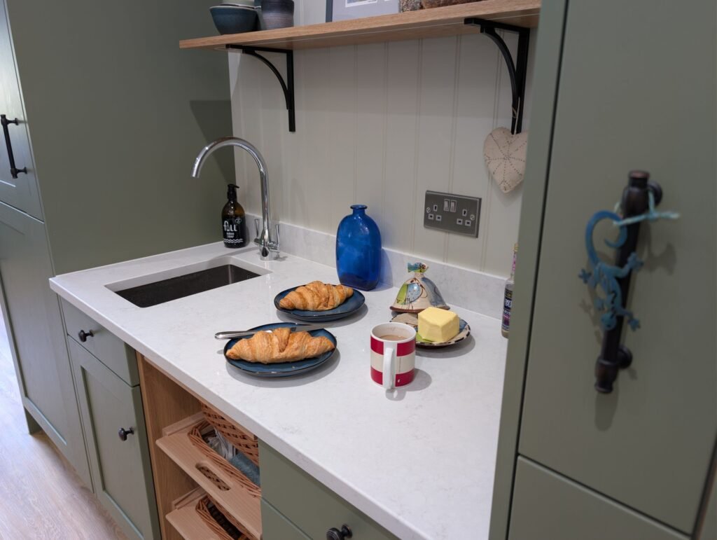 Small undermount sink in a Chalk Mist quartz worktop beside tall sage green units, with open shelves above