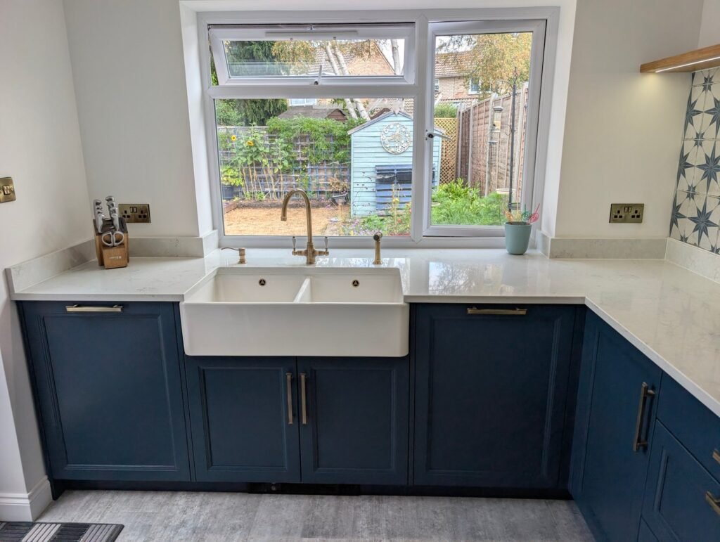 L-shaped Chalk Mist quartz worktop with Belfast sink and brass tap under a garden window in a navy shaker kitchen