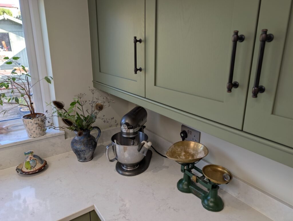 Sage green shaker wall units with black bar handles above a Chalk Mist quartz worktop and window sill