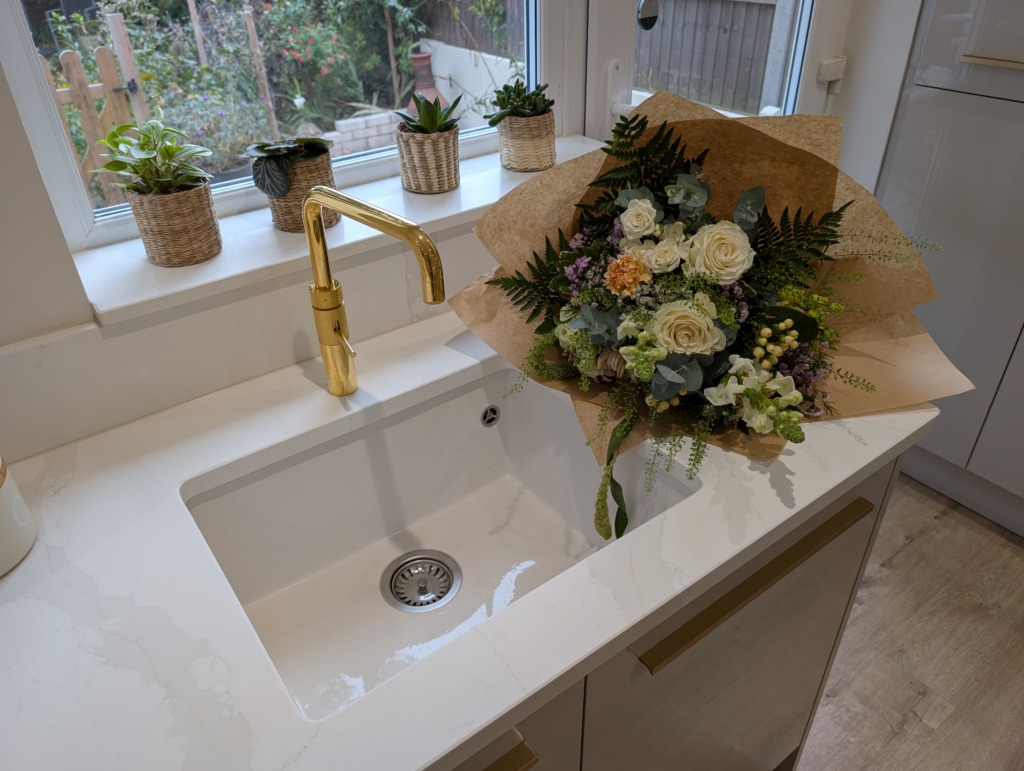Brass tap over a white undermount sink in a Gilded Vein quartz worktop, with flowers and plants on the quartz window sill