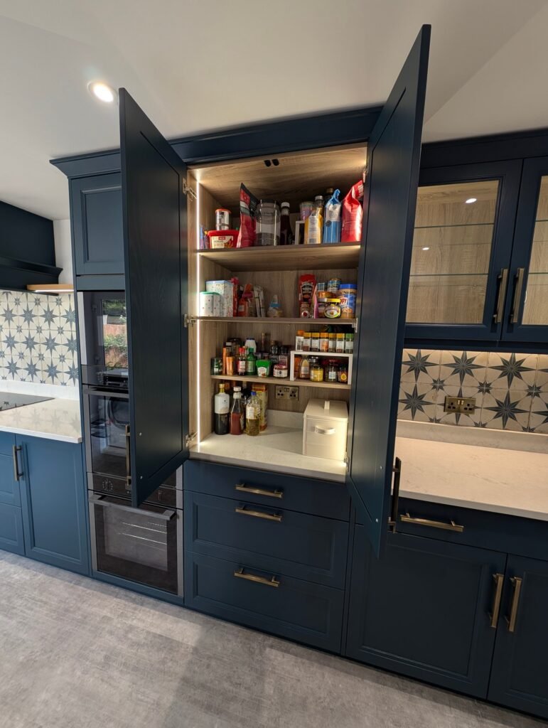 Navy shaker larder cupboard with doors open beside Chalk Mist quartz worktops and glazed wall units